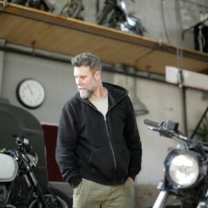 Confident middle aged male mechanic wearing workwear standing with hands in pockets and looking away while repairing motorbike in modern garage